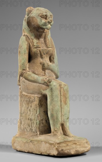Figurine of the goddess Uadjit with the head of a lioness; Uadjit mythology; Egyptian deities; lotuses; hieroglyphs; back pillars; thrones; Scen.Gal.Eg., 664; 30 BC – Late Period to Ptolemaic Period; Egypt; figurine; height 22 cm, width 11.5 cm, depth 7 cm; weight 1.024 kg., A seated anthropomorphic figure with a feline head and human body rendered in a weathered stone or ceramic material, showing a mix of pale green and beige tones with areas of darker brown wear; the figure wears a close-fitting garment and a wig-like head covering, with one hand resting on the lap and the other near the chest; the seat has a textured vertical pattern on its side and the base is rectangular with rounded edges, both showing similar greenish and beige coloration and signs of surface erosion; the overall background is a smooth gray.