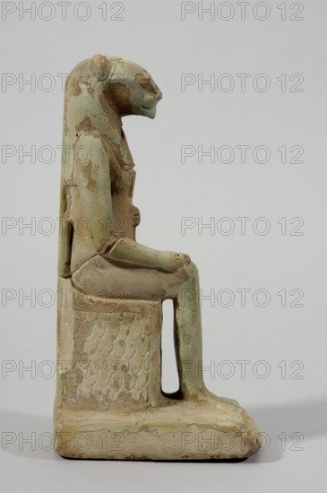 Figurine of the goddess Uadjit with the head of a lioness; maker, Uadjit mythology; lotuses; hieroglyphs; back pillars; throne; Late Period–Ptolemaic Period, 664–30 BC, Egypt; figurine; height 22 cm, width 11.5 cm, depth 7 cm, weight 1.024 kg., A seated statuette of an animal-headed figure shown in profile, made of a weathered material with areas of pale green and beige, depicts the figure wearing a long headdress that extends down the back, a defined arm resting on the lap, and legs bent at the knees with feet on a rectangular base; the seat has a textured pattern on its side, the surface shows patches of wear and discoloration in green and tan tones against a light neutral background.