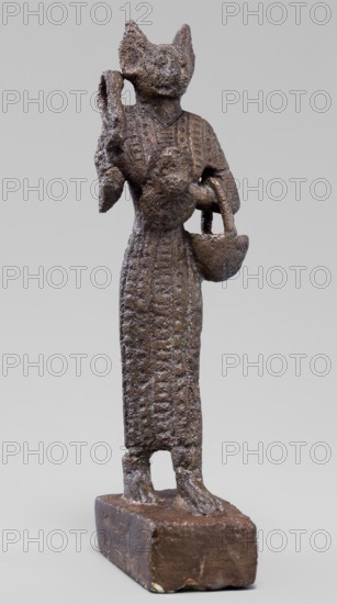 Figurine of the goddess Bastet. Figurine of a goddess with a cat's head, dressed in a tight-fitting, engraved robe. In her right hand she holds a bow sistrum, in her left an aegis. She also has a basket slung over her left forearm. Sistrum; aegis. Egyptian deities; Bastet mythology; Egyptian religion; Egyptian art. Ptolemaic Period, 332–30 BC. Figurine; height 9 cm, width 3.8 cm, depth 2 cm., A small textured statue with a rough, brown surface stands on a rectangular brown base; the figure has an elongated, vertical form wearing a long garment with patterned surface details and a pronounced chest area, and it holds a rounded object in one hand and a bowl-shaped object suspended from the other arm; the head is stylized with pointed ear-like projections and an indistinct facial surface, with the overall colors in the image being shades of brown on the figure and base against a pale gray background.