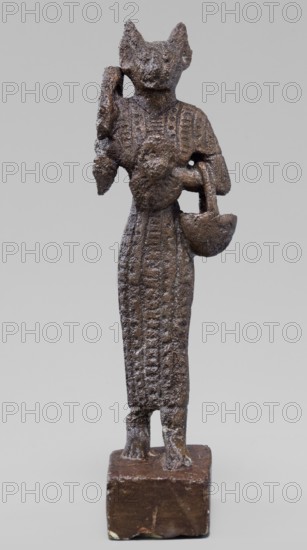 Figurine of the goddess Bastet. Figurine of a goddess with a cat's head, dressed in a tight-fitting, engraved robe. In her right hand she holds a bow sistrum, in her left an aegis. She also has a basket slung over her left forearm. Sistrum; aegis; Egyptian deities; Bastet mythology; Egyptian religion; Egyptian art. Ptolemaic Period, 332–30 BC. Egypt. Figurine, height 9 cm, width 3.8 cm, depth 2 cm., A small dark brown metallic figurine stands on a rectangular brown base, showing a slender humanlike body with a textured long garment and a head shaped like a feline with pointed ears; one arm is bent upward near the head while the other holds a rounded object that resembles a basin or bowl, and the surface displays a rough, pitted texture with variations of darker and lighter brown tones against a neutral gray background.