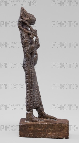 Figurine of the goddess Bastet. Figurine of a goddess with a cat's head, dressed in a tight-fitting, engraved robe. In her right hand she holds a bow sistrum, in her left an aegis, and she has a basket slung over her left forearm. Materials: sistrum, aegis. Subject: Egyptian deity Bastet. Date: 332–30 BC, Ptolemaic Period, Egypt. Type: figurine. Dimensions: height 9 cm, width 3.8 cm, depth 2 cm. Creator: unknown., The image shows a vertical sculpture of a standing anthropomorphic figure with an elongated body and textured surface, rendered in a dark brown material with rough, pitted areas and hints of lighter brown highlights; the figure stands on a rectangular base of similar dark brown color, with the feet positioned near the front edge; the head is stylized and angular with a pronounced snout-like projection, and a coiled element wraps around the upper torso and neck area; the arms are bent and held close to the chest, and the lower body features a patterned, ridged texture along th...