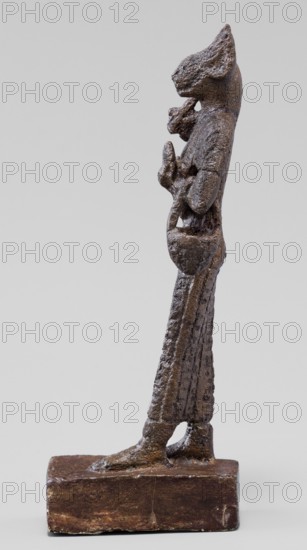 Figurine of the goddess Bastet. Figurine of a goddess with a cat's head, dressed in a tight-fitting, engraved robe. In her right hand she holds a bow sistrum; in her left an aegis. She also has a basket slung over her left forearm. Materials/function: sistrum, aegis. Egyptian deities, Bastet mythology. Egyptian religion, Egyptian art. Ptolemaic Period, 332–30 BC, Egypt. Figurine. Dimensions: height 9 cm, width 3.8 cm, depth 2 cm., A slender standing figurine with a rough textured surface in dark brown and rusty brown tones is shown in profile on a rectangular base of similar brown color; the figure has an elongated body and legs, a raised arm holding a small rounded object, and an animal-like head with pointed ears, with visible surface irregularities and worn areas.