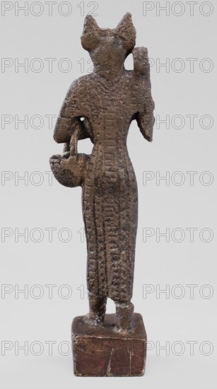 Figurine of the goddess Bastet. Figurine of a goddess with a cat's head, dressed in a tight-fitting, engraved robe. In her right hand she holds a bow sistrum, in her left an aegis, and she has a basket slung over her left forearm. Sistrum; aegis; Egyptian deities; Egyptian religion; Egyptian art. Ptolemaic Period, 332–30 BC. Figurine; height 9 cm, width 3.8 cm, depth 2 cm., A small standing figurine with a rough textured surface in shades of brown and dark brown, shown from the back on a square base that is a darker brown with lighter scuffs. The figure wears a long, vertically ridged garment extending to the ankles and has a narrow, elongated form. The head has two pointed protrusions and the shoulders are slightly rounded. One arm is bent and holds a rounded object with a handle-like element, while the other arm is raised with a bent elbow. The surface shows uneven pitting and small areas of lighter brown where the texture catches light. The background is a smooth pale gray.