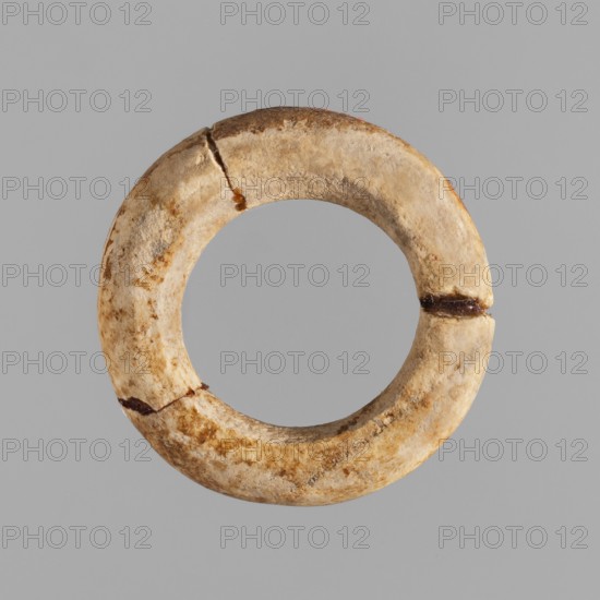 Ring — hair ornament; maker, Goluchów Greater Poland Voivodeship; rings; hair ornaments; ca. 1550–1069 BC; New Kingdom, Egypt; jewelry, A circular ring-shaped object with a textured, weathered surface showing shades of beige, cream, tan, brown, and rust; the object has an inner circular opening and a rounded outer edge, with visible cracks and a few darker brown spots or stains along its perimeter, set against a uniform light gray background.