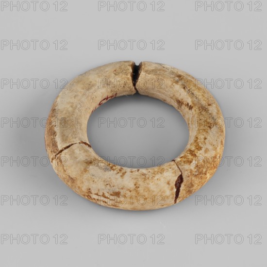 Ring - hair ornament, Goluchów Greater Poland Voivodeship; rings; hair ornaments; ca. 1550–1069 BC; New Kingdom, Egypt; jewelry, A circular ring-shaped object resting on a flat surface, showing a worn, uneven texture with visible cracks and breaks; the object has a predominantly beige and tan coloration with areas of darker brown mottling and some off-white patches; the surface appears slightly rough and aged, with a few darker brown fissures and a few small areas of reddish-brown along one inner edge; the background surface is smooth and light gray.