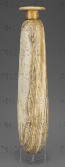 Alabastron, Slender alabastron with a distinct short cylindrical neck and a wide flat rim, provided with two tiny handles in the form of suppositories; vessel for oils and ointments, carved stone alabaster; Egyptian, Late Period, 664–332 BC; height 32 cm +1.5 cm base, width 5 cm, weight 1.11 kg., A tall, narrow elongated vessel with a rounded flat rim and short neck, displayed upright on a clear stand; the surface shows layered banding and streaks in shades of cream, beige, tan, and light brown with areas of off-white and subtle darker brown speckling, and smooth polished texture with natural irregularities and curved striations.