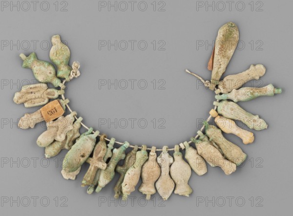 Fragment of a necklace of amulets 26 pieces, Goluchów Greater Poland Voivodeship; amulet necklace composed of 26 beige to greenish pieces including tit knots, ankh life signs and various objects; grave goods; ca. 1069–656 BC, Third Intermediate Period, Egypt; amulet, jewelry, A curved string holding a group of small pendant-like objects arranged like a garland, each pendant having a rounded, elongated, or slightly bulbous shape with small loops at their tops; the objects show a weathered, rough surface texture with areas of pitting and encrustation. The colors visible include shades of green, pale green, light tan, beige, off-white, and a few spots of darker brown; a small piece of twine or cord threads through the loops and appears off-white, and a short length of thicker twisted cord is tied near one end. The pendants vary slightly in shape and size and are spaced closely together along the cord, fanning outward to form a semicircular layout on a flat, smooth gray background.