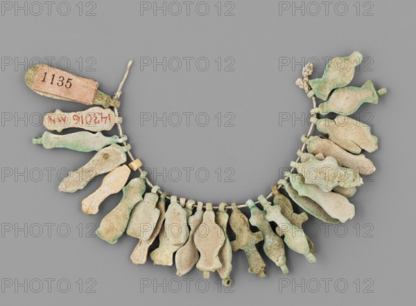 Fragment of a necklace of amulets 26 pieces, Goluchów Greater Poland Voivodeship, necklaces, grave goods, tit knots, ankh life signs, various objects, beige to greenish in color, ca. 1069–ca. 656 BC; Third Intermediate Period, Egypt, amulet, jewelry, A curved string with a group of small, flat, irregularly shaped pendants hanging from it, each pendant in varying pale colors including green, light beige, and muted turquoise with areas of discoloration and patina; the string appears off-white and knotted at one end, and a small rectangular tag is attached near one knot; the pendants vary slightly in shape and thickness and some show rough or flaked surfaces and darker green or brown spots against the otherwise pale tones.