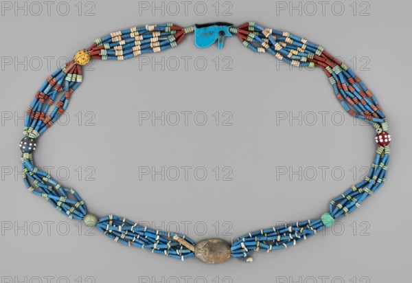 Necklace with a scarab and an udjat eye. Necklace composed of six rows of faience tubular and disc-shaped beads, divided into short sections by faience and glass beads. Two amulets complement the whole – a scarab and the eye of Horus udjat. c. 1650–1800 BC, Egypt. Jewelry amulet; height 70 cm., An oval arrangement of beaded strands on a gray background forming a necklace with a central oval bead in a metallic brown tone flanked by several strands of cylindrical blue beads interspersed with small off-white and pale green spacer beads, a few round beads in green and mottled turquoise, a dark bead with white dots, a red bead with white dots, a yellow round bead with small red marks, multiple short sections of reddish-brown tubular beads, and a small irregular bright blue clasp-like element; overall colors visible include blue, off-white, pale green, reddish-brown, yellow, red, dark brown, metallic brown, turquoise, and gray.