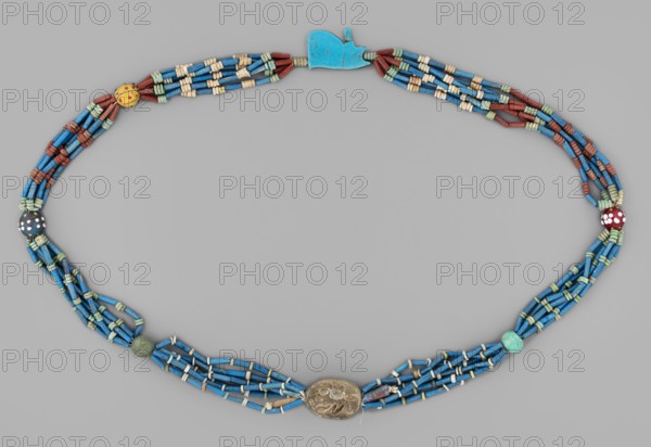 Necklace with a scarab and an udjat eye. Necklace composed of six rows of faience tubular and disc-shaped beads, divided into short sections by faience and glass beads. Two amulets complement the whole – a scarab and the eye of Horus udjat. c. 1650–1800 BC, Egypt. Jewelry/amulet, height 70 cm., An oval beaded necklace arranged on a neutral gray background composed of several strands of tubular blue beads interspersed with small cylindrical beads in pale green, cream, and reddish-brown, featuring a few round accent beads in yellow with red dots, black with white dots, red with white dots, a mottled green bead, and a turquoise bead, and a flat turquoise clasp piece and an oval metallic pendant with textured surface.
