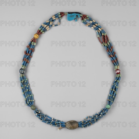 Necklace with a scarab and an udjat eye. Necklace composed of six rows of faience tubular and disc-shaped beads, divided into short sections by faience and glass beads. Two amulets – a scarab and the eye of Horus udjat. c. 1650–1800 BC, Egypt. Jewelry/amulet, height 70 cm., A beaded necklace arranged in an oval shape on a neutral background, composed of several strands of small cylindrical beads primarily in shades of blue, interspersed with a few beads in green, cream, red, yellow, and brown; there are a few larger round and oblong beads placed at intervals, including a dark metallic oblong bead and a few round beads in green, yellow, red, and dark blue with spotted patterns; the strands are grouped and tied together with small cream and green spacer beads and the closure area includes a flat blue component.