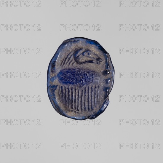 Scarab with a ram's head, ram's head ornament, sacred animal protective amulet in navy blue glass; scarab amulet, height 2.4 cm, width 1.9 cm; 664–525 BC, Late Period, 26th Dynasty, Egypt;, A small oval object with irregular edges photographed against a plain light gray background; the object has a textured surface in shades of deep blue and metallic gray, with a darker blue central oval area and vertical striations beneath it, and a rounded raised form toward the top that suggests a curved shape.