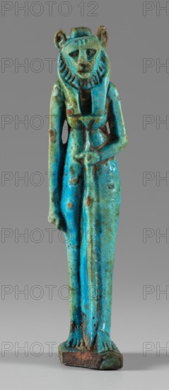 Amulet — figurine of a lion-headed goddess. A lion-headed goddess, in a tripartite wig and a narrow robe, walks with a lotus scepter in her left hand the right one is lowered. Presumably the goddess Sekhmet; possibly Wadjit or Bastet. Late Period, Egypt, 664–332 BC. Amulet; height 8.8 cm, width 1.7 cm., A small standing figurine shown full-length against a neutral background, with a stylized human body and an animal-like head, wearing a striped headdress and a long garment; the figure's posture is straight with one arm hanging down and the other bent across the torso holding a long object. The surface shows a weathered, glazed appearance with predominant turquoise and blue tones, along with areas of greenish patina and patches of brownish discoloration and darker spots. The base is rectangular and has worn brown and green coloration. The overall finish is glossy in places and uneven, with visible texture variations and small pits.