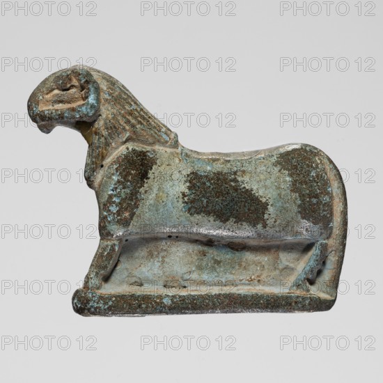 Amulet — ram;, Ram-shaped protective amulet, sacred animal motif. Late Period, Egypt, 664–332 BC. Materials not specified. Dimensions: height 2.2 cm, width 0.7 cm., A small sculptural object shaped like a stylized animal with a mane, shown in profile and resting on a rectangular base; the surface has an uneven, weathered texture with areas of roughness and pitting. The visible colors include greenish-blue patina, brownish rust tones, and pale beige areas where the surface is worn. The animal form has a pronounced head with a carved mane, a rounded back, and simplified legs merged into the base, with shadows and highlights emphasizing the contours and the aged surface.