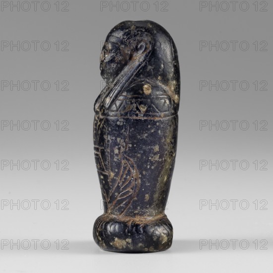 Amulet — god Osiris in hydria; maker, Osiris canopic figure; Egyptian deities, Egyptian religion; 1st–2nd century AD Roman period, Egypt; amulet; height 4.9 cm, width 2.1 cm, A small carved figure shown in profile standing on a rounded base, rendered in dark bluish-black stone with areas of lighter brownish and greenish surface wear; the figure has a rounded head with a smooth, elongated headdress or hair extending down the back, a defined forehead and nose, and a slightly protruding chin; the torso is decorated with incised geometric and linear patterns, including triangular and banded motifs, and the lower body shows carved winglike and feathered designs; surface pitting and small patches of lighter discoloration are visible across the object, and the background is a smooth neutral gray.