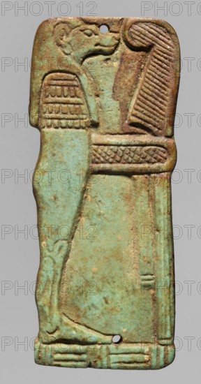 Amulet - plaque with a depiction of the god Hapi. Relief figurine of the god Hapi, shown in right profile, in the form of a mummy with the head of a baboon in a wig. In front of the figure is an ostrich feather on a banner, connected to the god's body with a piece of decorative material. Mat under the feet. Presumably an element of a mummy net. Late Period, 26th Dynasty, Egypt, 664–525 BC. Amulet; height 6.2 cm, width 2.6 cm., A vertical rectangular object with rounded corners shown against a gray background, featuring a relief of a standing figure with a stylized animal head and a long dress; the surface shows a mix of turquoise, green, brown, and tan tones with areas of darker brown patina and lighter worn spots, and a few small holes near the top and bottom; decorative carved patterns appear on the figure's headdress, collar, and a horizontal band, and there are several linear incised details along the right edge and bottom.