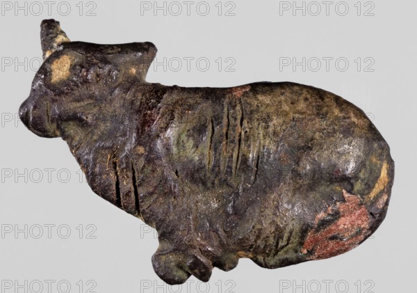 Amulet - bound sacrificial bull, Goluchów Greater Poland Voivodeship; bulls, sacrifices; Scen.Gal.Eg., sacrifice; 332–30 BC, Ptolemaic Period, Egypt; amulet; height 3.3 cm, width 2 cm, A small sculptural object shaped like an animal is shown against a plain light gray background, with a rough, textured surface that appears weathered and irregular. The object displays a mix of dark brown and black tones with areas of greenish patina and patches of muted yellowish-beige and reddish-brown. The form includes a rounded body, a defined head with a snout and ear-like projection, and a small protrusion beneath suggesting a leg or paw; the surface has several shallow grooves and cracks and spots where the original surface is worn away. There are localized areas of slight sheen and scattered lighter flecks that contrast with the overall darker coloration.
