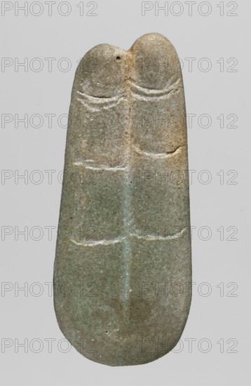 Amulet — two fingers; maker, Goluchów Greater Poland Voivodeship; fingers; amulets — body parts; grave equipment; 664–525 BC; Late Period, 26th Dynasty, Egypt; amulet; height 5.5 cm; width 2.4 cm, A single elongated, rounded object resembling a carved stone is centered against a plain light gray background, showing a muted palette of greenish-gray with areas of beige and brown. The stone has a smooth surface with subtle weathering and speckled texture, and a vertical ridge runs down its middle dividing the piece into two symmetrical lobes at the top. Shallow, curved incisions form parallel horizontal lines across the upper and middle sections on both sides of the central ridge, and a small circular hole is visible near the top left lobe. The overall shape tapers slightly toward the top and rounds more at the bottom, and the stone displays variations of color from pale green to sandy tan with faint lighter highlights along the carved grooves.