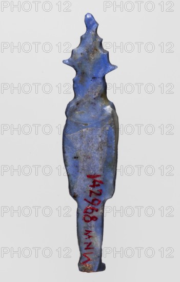 Amulet — Nehebkau; maker, Nehebkau myth., Egyptian deities, Egyptian religion; dark blue glass; Ptolemaic Period, 332–30 BC, Egypt; amulet; height 4.7 cm, width 1 cm., A single elongated object with an irregular pointed top and a tapered bottom, shown against a plain light gray background. The object is primarily shades of blue with areas of darker blue, lighter blue, and some greenish-blue discoloration. There are scattered brownish spots and patches of dirt or staining across the surface, and a small area near the bottom shows a rusty orange color. The surface appears worn and slightly pitted with uneven texture, and the silhouette includes a few protruding lobed shapes near the upper portion.