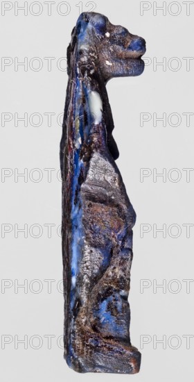 Amulet — goddess Toeris;, Dark blue glass. Ptolemaic Period, 332–30 BC, Egypt. Amulet. Height 4.4 cm; width 1 cm., A narrow vertical object resembling a carved figurine is shown against a plain light gray background; the object has an irregular textured surface with a mix of dark blue, deep brown, black, and lighter bluish-gray areas, along with a few small pale whitish patches; it appears elongated with a raised head-like shape at the top and subtle contours suggesting limbs or facial features, and the surface shows glossy and matte areas with some rough, granular patches and streaks of color running along its length.