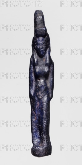 Amulet - standing goddess Isis, Isis myth., Egyptian deity goddess, navy blue glass, 332–30 BC Ptolemaic Period, Egypt, amulet, height 5.1 cm, width 3.9 cm, A slender vertical figurine with a slightly textured surface rendered in deep blue with areas of darker navy and small patches of lighter blue and brownish wear, showing a simplified human-like form with a narrow head, shoulders, and elongated torso, subtle raised shapes suggesting a face and chest, short arm-like projections along the sides, and a tapered lower body ending in a rounded base, photographed against a plain pale gray background.