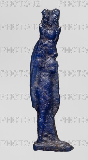 Amulet - goddess Hathor; Dark blue glass with a touch of violet; Ptolemaic Period, 332–30 BC, Egypt; amulet; height 5.4 cm, width 1.1 cm; depicts Hathor Egyptian goddess., A slender vertical object shaped like a stylized standing figure with an elongated body and a suggestion of shoulders and hips, rendered in a deep blue color with patches of darker and lighter blue and small areas of brownish discoloration, showing a rough, uneven surface texture with visible bumps and irregularities, standing on a narrow flat base that is also blue with worn edges, all set against a plain light gray background.