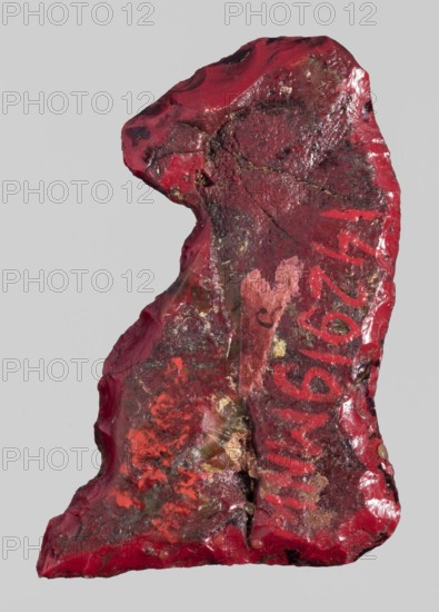 Amulet — sitting baboon; red glass; Ptolemaic Period, 332–30 BC, Egypt; amulet; height 3.2 cm, width 2.6 cm; subjects: baboons, sacred animals, Egyptian religion., A rough, irregularly shaped object with a glossy surface in shades of red, dark red, and maroon, showing areas of brighter red and patches of muted pink and coral; a few small spots of tan and pale yellow are visible within textured, uneven surfaces, along with areas of darker brown and black mottling; the object has jagged edges, concave and convex surfaces, and visible chips and fractures, set against a plain light gray background.