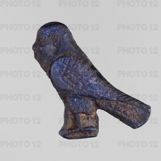 Amulet - Ba, Ba myth., Egyptian religion, grave goods, navy blue glass, 332–30 BC; Ptolemaic Period, Egypt, amulet, height 2.7 cm, width 3.1 cm, A small sculpted object resembling a bird shown in profile with a rounded head, short beak, compact body, and a long tapered tail; the surface appears textured and worn with a mottled combination of deep blue and brown tones, with a few lighter spots and areas of darker shadow; the bird stands on a small rectangular base of similar rough texture and color against a plain light gray background.