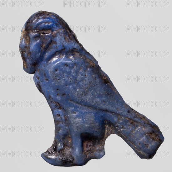 Amulet — Ba figure;, Egyptian Ptolemaic Period 332–30 BC. Navy blue glass amulet representing the Ba myth. from Egyptian religion; used as grave goods. Height 3.2 cm, width 3.8 cm., A small worn figurine shaped like a crouching animal with a rounded head and pronounced beak-like snout, mostly covered in a vibrant blue surface with areas of darker brown and black discoloration and pitting; the form shows smooth, rounded contours with a suggestion of a hunched back and a short tail-like projection, and the lower area appears rougher with patches of darker brown and black on the underside and along some edges.