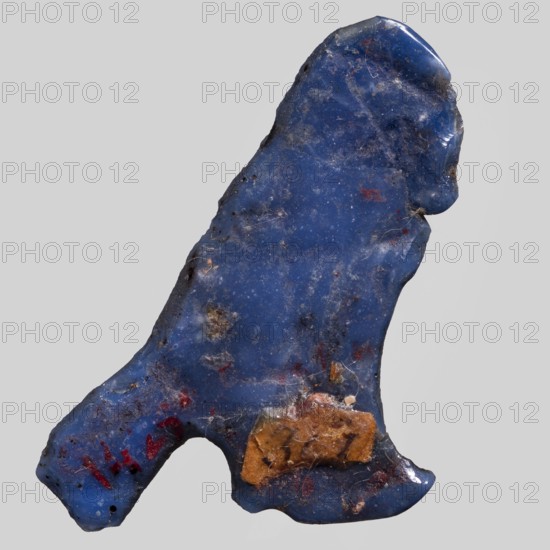 Amulet — Ba, Ba myth., Egyptian religion; grave good; navy-blue glass; Ptolemaic Period, 332–30 BC, Egypt; amulet; height 3.2 cm, width 3.8 cm, A roughly irregular, roughly triangular stone-like object with a glossy surface predominantly in deep blue, showing patches of lighter blue and tiny pale speckles; there are several small areas of brown and tan material adhered near one edge, a few reddish spots scattered across the surface, and some thin fibrous debris clinging in places; the object has uneven edges with a few rounded protrusions and indentations, and the background is a uniform pale gray.