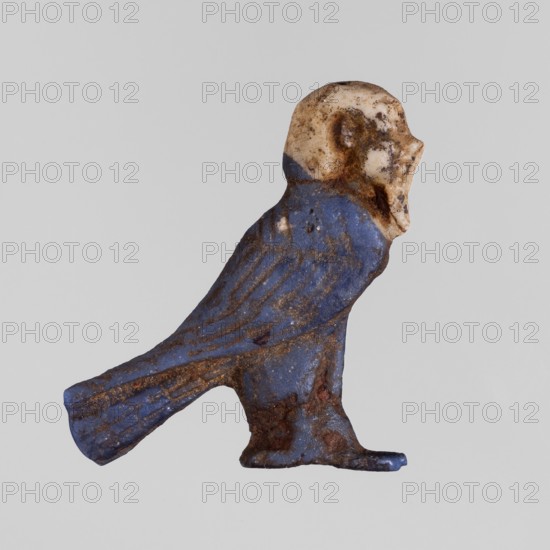Amulet - Ba, Ba myth., Egyptian religion, grave goods, white human head on navy blue bird background, Ptolemaic Period 332–30 BC, Egypt, amulet, height 2.6 cm, width 3 cm, A small sculpted figure viewed in profile showing a birdlike body with a rounded head, carved wings and a tail, resting on a short base. The surface displays a mix of blue and cream tones with areas of brown discoloration and darker brown spots; the blue covers most of the body and tail while the cream is concentrated on the head. The texture appears worn and irregular with patches of roughness and some pitted areas, and the overall shape is compact with a slightly curved posture.
