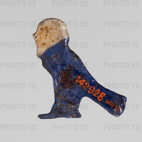 Amulet - Ba, ba myth., Egyptian religion; grave good: white human head on navy blue bird background;, amulet; date: 332–30 BC, Ptolemaic Period, Egypt; dimensions: height 2.6 cm, width 3 cm., A small irregular object shaped like a stylized figure with a rounded top and a protruding leg, showing a mix of colors including blue, white, brown, orange, and a few dark rusty spots, with a glossy surface on parts and a matte, speckled appearance on the white area, and some rough textured areas on the blue portion.