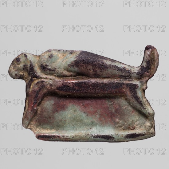 Amulet - mummy lying on a lion bed; mummification bed iconography, mummies, lions; amulet, height 2.2 cm, width 3.5 cm; 332–30 BC, Ptolemaic Period, Egypt., A small sculptural object showing a stylized animal figure reclining on a rectangular base, rendered with rounded, simplified forms and a curled tail; the surface shows areas of green, pale blue, reddish-brown, dark brown, and spots of black, with worn and pitted texture and traces of encrustation; the background is a plain light gray.