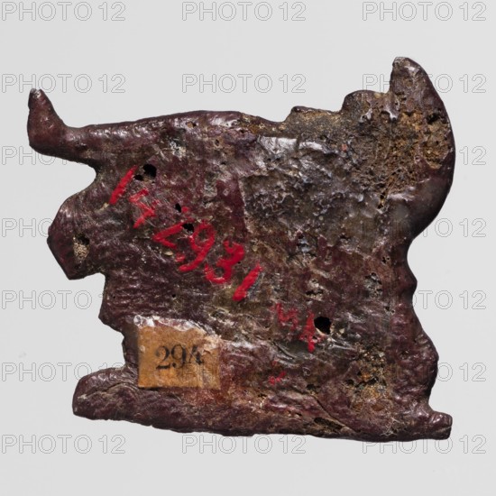 Amulet - mummy lying on a lion-shaped bed; maker, mummification bed iconography; subjects: mummies, lions; material: dark red glass; date: 332–30 BC, Ptolemaic Period, Egypt; function: amulet; dimensions: height 2.6 cm, width 2.9 cm., The image shows an irregularly shaped, flat object with a rough, pitted surface and uneven edges, appearing worn and corroded; the dominant colors visible are dark brown and deep reddish-brown with areas of lighter brown and patches of yellowish-brown, along with small accents of bright red paint and some tiny darker spots; the background is a uniform light gray.