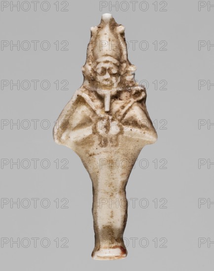 Amulet — standing god Osiris; light yellow glass; Ptolemaic Period, 332–30 BC, Egypt; amulet; height 3.6 cm, width 1.6 cm; Egyptian deities; Osiris; Egyptian religion, A small carved figure in off-white and beige tones with brownish soil or age discoloration throughout, showing a stylized human shape with a pointed headdress, a rounded face area, and a suggested torso with arms folded or held together; the surface has rough and smooth areas with darker brown flecks and smudges, and a single small area of reddish-brown near the bottom; the background is a flat light gray.