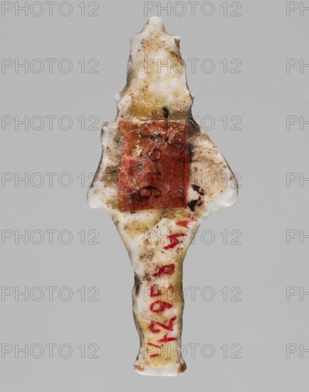 Amulet - standing god Osiris; Light yellow glass; 332–30 BC, Ptolemaic Period, Egypt; amulet; height 3.6 cm, width 1.6 cm; Egyptian deities; Osiris; Egyptian religion, An elongated, irregular object with a central narrow shaft and flared, wing-like projections near the top, appearing to be made of a hard, glossy material with a worn and dirty surface; the object shows a mix of off-white and yellowed areas with brownish grime and smudges, and a rectangular patch of muted reddish-brown material adhered near the upper middle, with traces of darker brown spots and fine fibrous debris around it; there are also several small red marks along the lower portion of the shaft, and the background is a uniform light gray.
