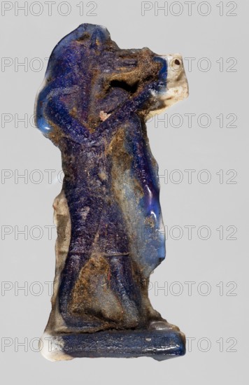 Amulet — a deity with a jackal's head Anubis, dark blue glass; Ptolemaic Period, 332–30 BC, Egypt; amulet; height 3.3 cm, width 1.5 cm., A small carved figure with a rough textured surface, predominantly deep blue with patches of lighter blue and areas of brownish dirt-like deposits, showing traces of white at the base and near the head; the figure appears upright with a hooded or rounded head shape and an arm bent across the chest, a base supporting the figure, and irregular worn edges and pitted spots across its surface.