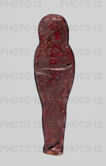Amulet — mummy-shaped figurine, mummy-shaped figurines, grave goods; back smooth. Dark red glass. 332–30 BC; Ptolemaic Period, Egypt. Amulet. Height 3.8 cm, width 1 cm. /author., A single elongated object shaped like a simplified human figure fills the center of the image, with a rounded head and a tapered lower body, photographed against a plain light gray background. The object's surface is worn and uneven, showing a predominant dark red color with patches of deeper maroon and small areas of brown and black. The top and edges display a few rough spots and tiny pits, and there are subtle variations in sheen where the surface is smoother or scuffed. The lower end appears slightly creased and shows a small exposed area of tan.