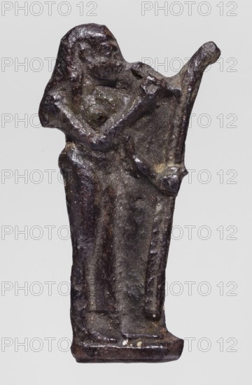 Amulet - goddess Seshat; maker, Seshat mythology; Egyptian deities; Egyptian religion; goddesses; harp iconography; dark brown glass; 332–30 BC, Ptolemaic Period, Egypt; amulet; height 3.4 cm, width 1.7 cm, A small dark metallic figurine showing a standing humanlike figure with a rounded head and simplified facial features, a raised arm bent at the elbow, and another arm extending upward and outward; the body appears draped in a long robe with visible folds and a slightly flared base, and the surface shows an uneven, pitted texture with areas of lighter and darker metallic tones including dark brown, black, and hints of gray and greenish patina.
