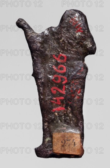 Amulet — goddess Seshat;, Dark brown glass; Ptolemaic Period, 332–30 BC, Egypt. Depicts Seshat Egyptian goddess associated with writing, harp iconography. Amulet; height 3.4 cm, width 1.7 cm., A roughly rectangular, irregularly shaped metal object with a rough, pitted surface is shown against a plain light gray background; the object has a dark brown to nearly black patina with areas of metallic sheen and hints of greenish corrosion, and there are a few strokes of bright red paint on its surface; the top edge is uneven with a protruding curved nub on one side and a rounded raised area on the other, the lower edge flares slightly and has a small attached rectangular fragment of brownish material with a worn appearance.