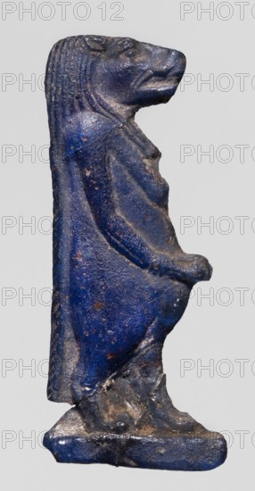 Amulet - goddess Toeris; Dark blue glass with a violet tint; Ptolemaic Period, 332–30 BC, Egypt; amulet; height 4 cm, width 1.6 cm; Egyptian deity/goddess, Toeris., A small sculpted figure standing upright, shown in profile, with a head shaped like an animal and a draped garment that covers the body; the surface appears textured and worn with uneven edges and small pits, and the figure stands on a rectangular base. The visible colors include shades of deep blue, darker blue, and areas of brownish discoloration, with small spots where the underlying material shows through.