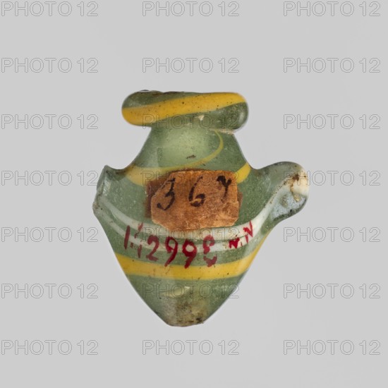 Amulet - heart ib, amulet, hearts ib representing a heart, amulet of a body part, grave equipment, New Kingdom, 18th Dynasty, ca. 1550–1295 BC, Egypt; height 2.2 cm, width 1.6 cm, A small glass object with a translucent green body and a few curved yellow bands, shaped like a tiny vessel with a rounded top and a wider flared midsection, showing some chips and rough edges and a small brown paper fragment stuck on one side; the surface has a few darker spots of dirt and wear and the colors visible are green, yellow, brown, white, and hints of red.