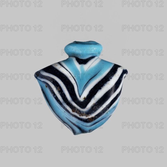 Amulet in the shape of a heart ib, made of blue glass decorated with parallel lines forming a zigzag on the front and back in navy blue and white. The amulet is vertically pierced, the hole framed at the top with a blue ring, with residual handles on the sides. c. 1550–1295 BC; New Kingdom, 18th Dynasty, Egypt. Amulet heart/ib, height 1.9 cm, width 1.9 cm., A small object with a shield-like shape centered on a plain light gray background, showing layered curved bands in bright turquoise, deep black, and white that form a chevron pattern, with a rounded turquoise top section; the surface shows tiny spots of brown discoloration and subtle texture variations, and the overall colors visible are turquoise, black, white, brown, and light gray.
