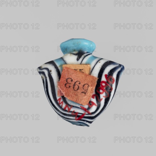 Amulet in the shape of a heart ib, made of blue glass decorated with parallel lines forming a zigzag on the front and back in navy blue and white. The amulet is vertically pierced; the hole is framed at the top with a blue ring, and there are residual handles on the sides. c. 1550–1295 BC; New Kingdom, 18th Dynasty, Egypt. Amulet heart ib, height 1.9 cm, width 1.9 cm., A small decorative object with a rounded top in light blue sits against a plain gray background; the body of the object shows alternating curved bands of black and white surrounding a central area with blue and white tones, and there is a piece of rough reddish-brown material attached to the front partially covering the central area; there are also a few small red markings on the lower right side of the object and some tiny dark speckles across the surface.