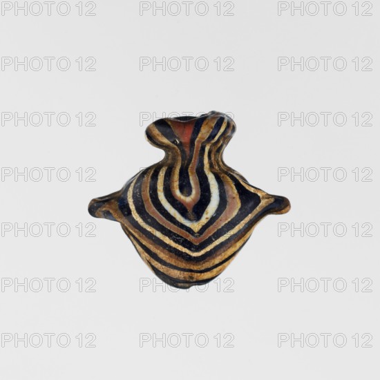 Amulet in the shape of a heart ib, made of black and brown glass decorated with parallel lines forming a zigzag on the front and back in beige and white. The amulet is vertically pierced; the hole is framed at the top with a beige ring, and there are residual handles on the sides. c. 1550–1295 BC; New Kingdom, 18th Dynasty, Egypt. Amulet; height 1.6 cm, width 1.7 cm., A small object with a rounded, bulbous shape and a narrow neck, displaying concentric, wavy bands in black, brown, tan, white, and reddish tones; the surface appears smooth and slightly glossy, with the bands forming a symmetrical, layered pattern that follows the contours of the form, and small protruding lobes on either side.