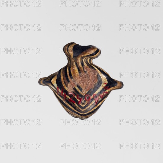 Amulet in the shape of a heart ib, made of black and brown glass decorated with parallel lines forming a zigzag on the front and back in beige and white. The amulet is vertically pierced; the hole is framed at the top with a beige ring, and there are residual handles on the sides. c. 1550–1295 BC; New Kingdom, 18th Dynasty, Egypt. Amulet; height 1.6 cm, width 1.7 cm., A small object with an organic, irregular shape sits against a plain light gray background; the object has layered, flowing stripes in black and tan/golden tones with areas of brown and a few spots of red, and its silhouette suggests a rounded central form with two short protrusions on opposite sides and a narrowed neck at the top.