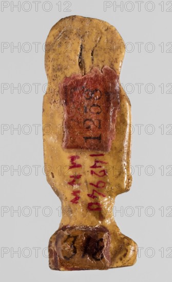 Amulet - tit, tit knots, protective amulet, grave good, with hieroglyphs, yellow glass; Ptolemaic Period, 332–30 BC, Egypt; amulet, height 3.3 cm, width 1.2 cm., A small carved object resembling a stylized human figure viewed from the back, with a smooth, worn surface in a yellow-beige color mottled with brown spots and darker speckles, a reddened rectangular patch near the upper back area, a few strokes of red pigment along the spine area, and a darker brown rounded base area; the background is a neutral light gray.