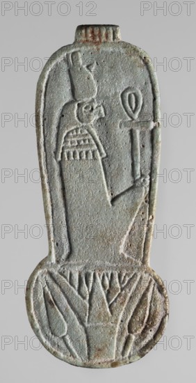 Amulet – weight of a menit necklace, An amulet in the shape of the counterweight of a menit necklace, with the image of a mummy-shaped, falcon-headed deity in a double crown holding an ankh in the longitudinal part and a lotus flower with two buds in the round lower part. On the reverse is a column of text with a sacrificial formula to the goddess Uadjit. Materials/icons: Re mythology, Uadjit, double crown, ankh, lotuses, hieroglyphs, sacrificial formulas, Egyptian deities. Date: 664–332 BC, Late Period, Egypt. Type: amulet. Dimensions: height 9.2 cm, width 3.3 cm., A vertical carved object with a rounded lower end, showing a raised profile of a human figure wearing a tall headdress and a patterned collar, holding a staff-like item; the surface is weathered and pitted with textured carving lines. The piece displays a palette of pale greenish-blue with areas of gray and small patches of brownish rust and darker green, with subtle variations across the relief and edges. The lower rounded...