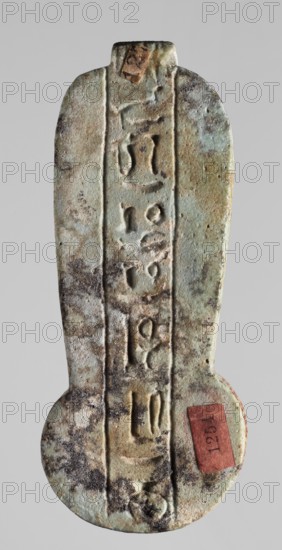 Amulet – weight of a menit necklace;, An amulet in the shape of the counterweight of a menit necklace, with the image of a mummy-shaped, falcon-headed deity in a double crown holding an ankh in the longitudinal part and a lotus flower with two buds in the round lower part. On the reverse is a column of text with a sacrificial formula to the goddess Uadjit. Materials and iconography include Re, lotuses, menitoses, Uadjit, double crown, Egyptian deities, hieroglyphs, ankh, sacrificial formulas; function: amulet. Late Period, Egypt, 664–332 BC. Height 9.2 cm; width 3.3 cm., A vertically oriented weathered object with a rounded bottom and a narrower rounded top, displaying a rough, pitted surface and carved linear grooves running down its center. The surface shows a mottled mix of green, pale green, brown, tan, gray, and black tones with areas of darker spotting and lighter patches. There are a few rectangular paper fragments affixed near the top and lower right, one of which is reddish in...