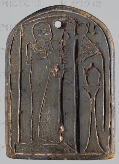 Amulet - stele with a depiction of the god Ptah and a falcon on the reverse;, Ptah mythology; falcon iconography; naos, plaques; Egyptian deities; 664–332 BC, Late Period, Egypt; amulet, stele; height 7 cm, width 5 cm, A carved rectangular plaque with a rounded top, primarily dark gray with areas of brownish wear, shows an incised profile of a standing human figure on the left and a tall stylized plant or object on the right; the figure is outlined in a lighter brownish tone and wears a short wig or headdress, a collar, and a long robe, holding a vertical staff-like object; there is a small round hole near the top center, and several thin incised borders frame the composition; overall the surface shows worn brown highlights against the dark gray background.